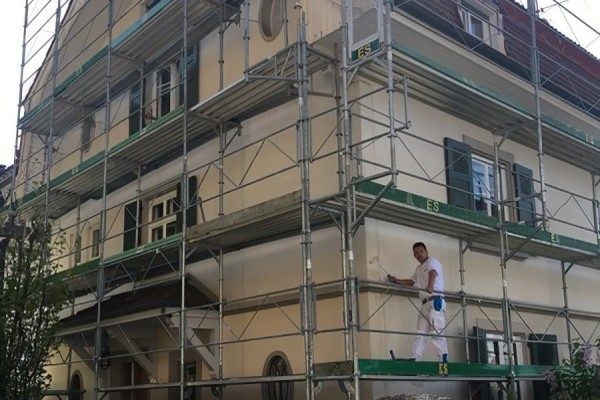 Maison ancienne entièrement échafaudée, un peintre au travail sur le plancher de l’échafaudage