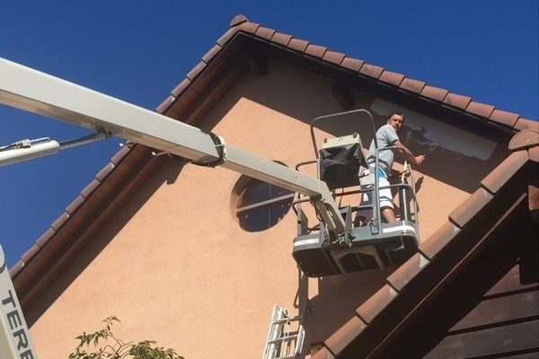 Peintre dans une nacelle élévatrice, sous le pignon et l’avant-toit d’une maison