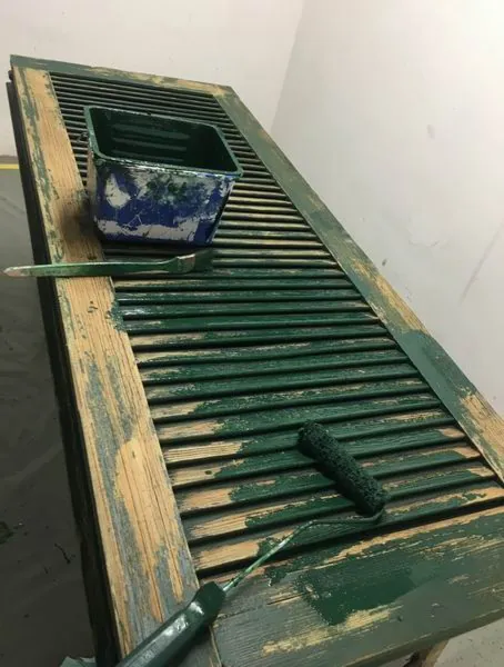 Volet à persienne posé sur l’établi, moitié gauche décapée jusqu’au bois, moitié droite repeinte en vert